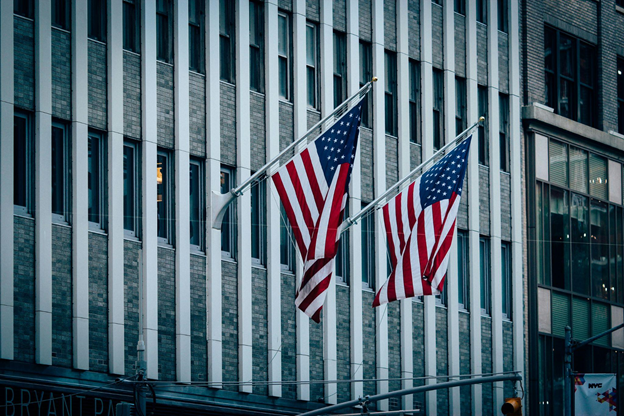 Two American flags wave in unison against a gray, vertical-lined building.